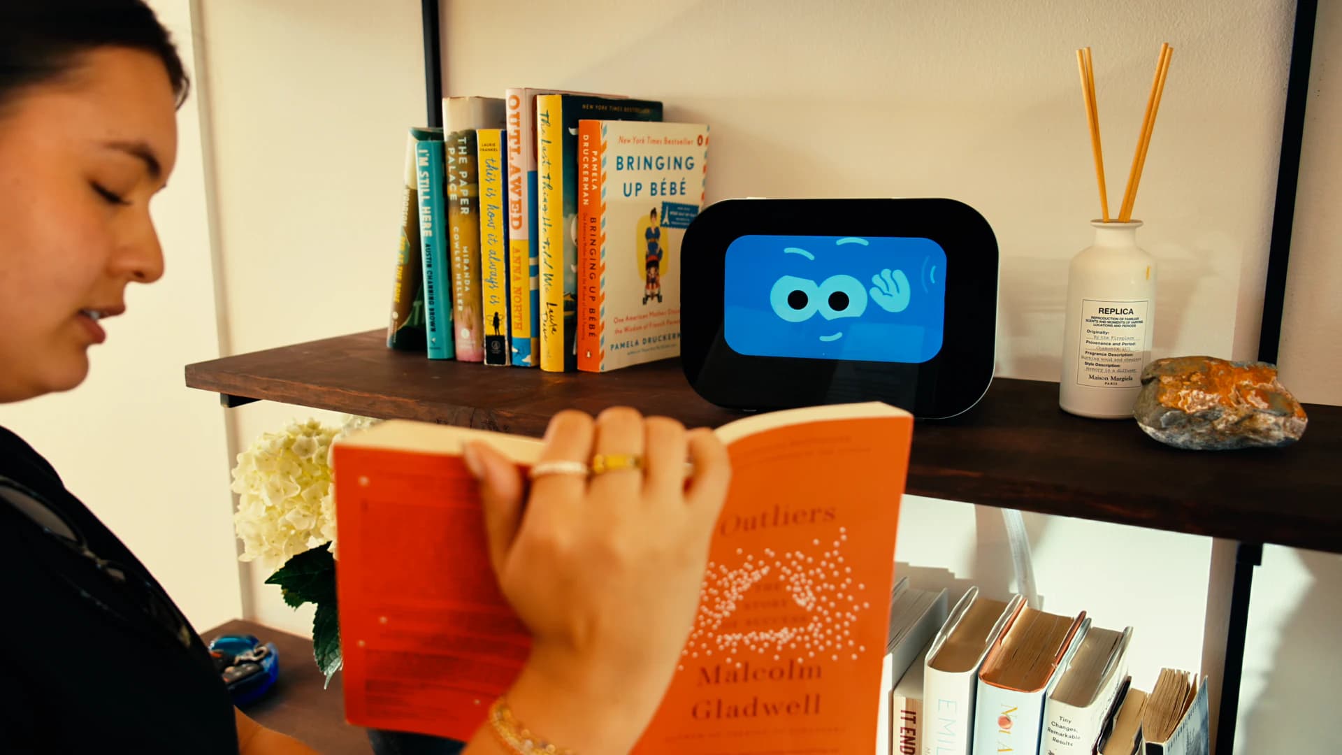 Rhem AI health robot on a bookshelf greeting with a friendly waving face while a woman reads nearby.