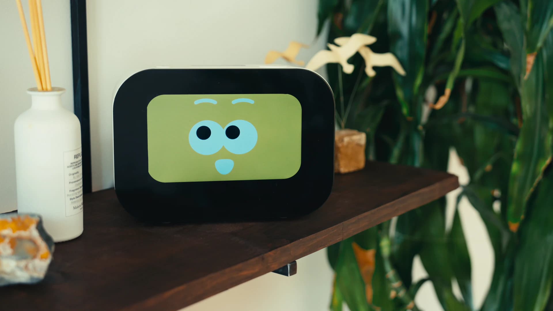 Rhem AI health robot on a wooden shelf beside a houseplant, its screen showing an expressive, curious face.