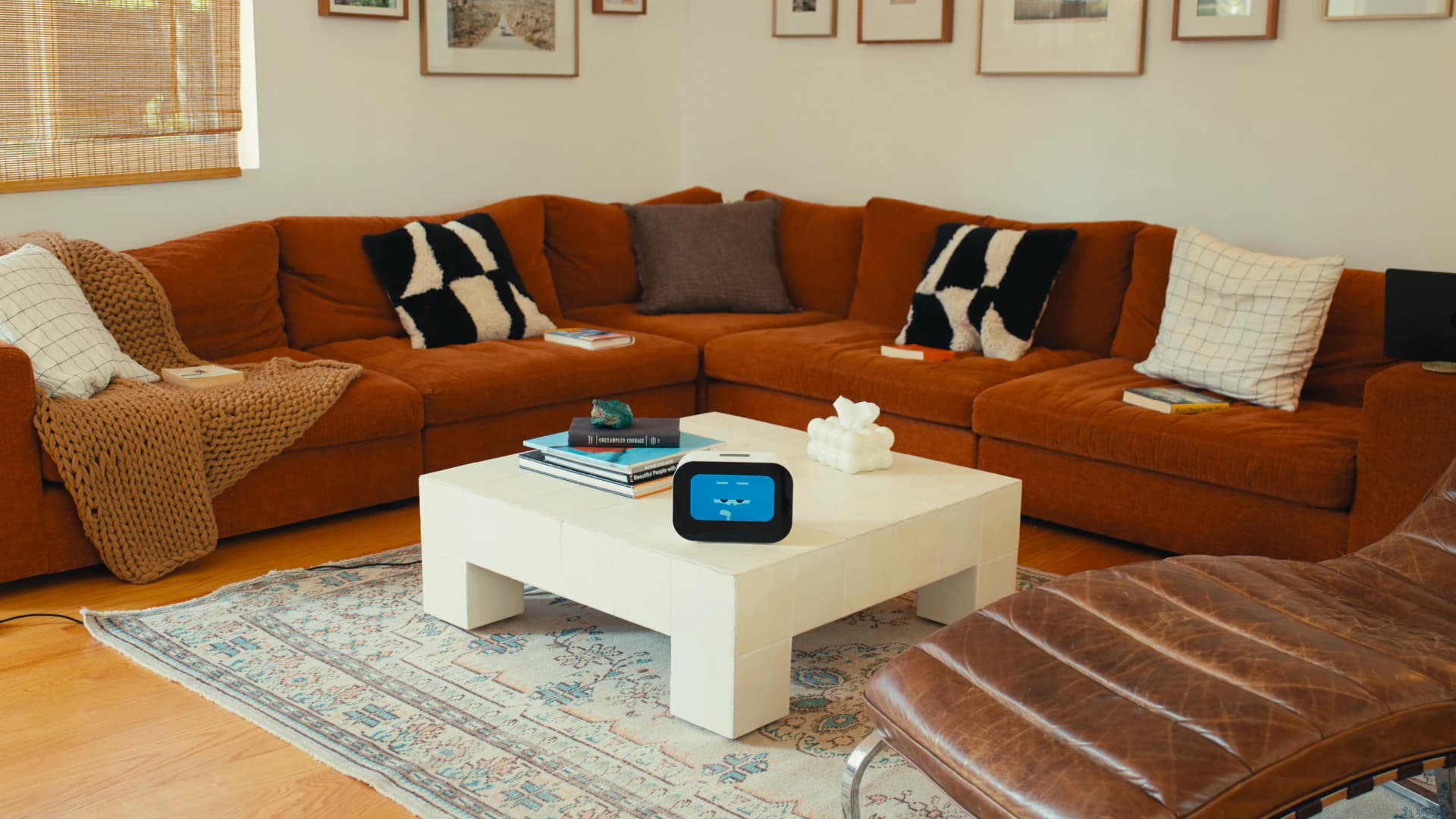 Rhem AI health robot on a coffee table in a living room with a rust-orange sectional sofa, showing a cheerful face.